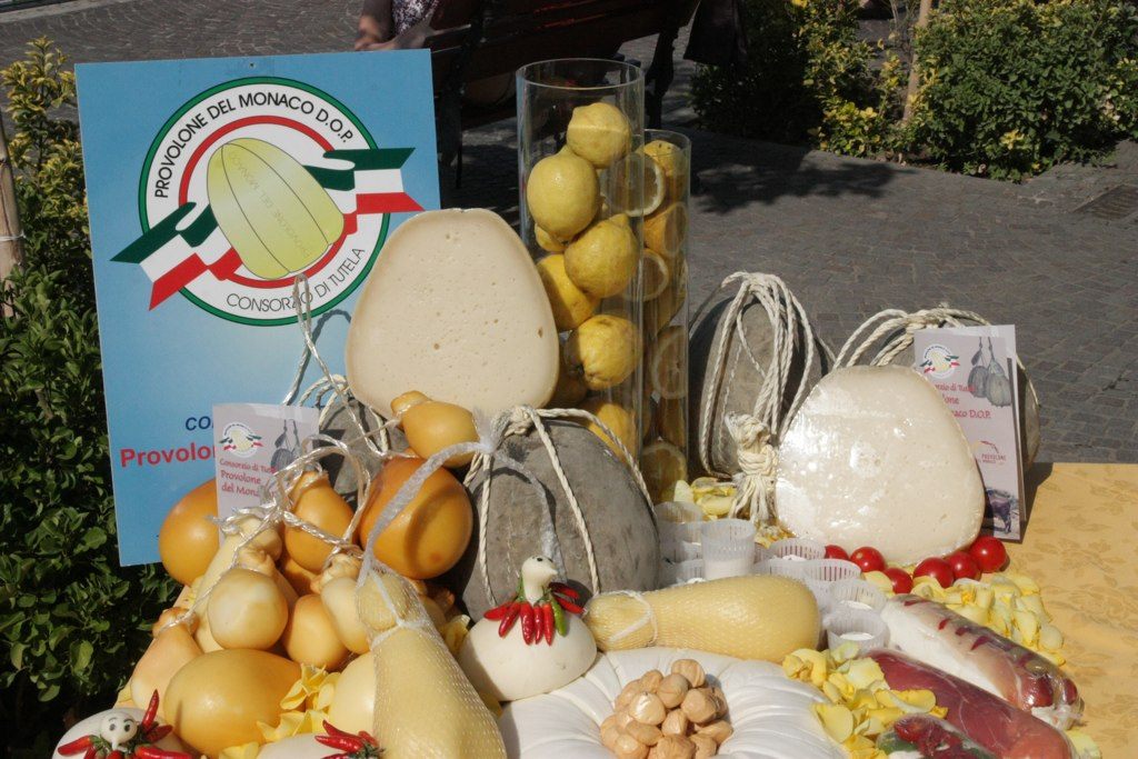 Forme di Provolone del Monaco DOP, intere e tagliate, legate con la corda ed esposte con altri latticini davanti al cartello del Consorzio di tutela